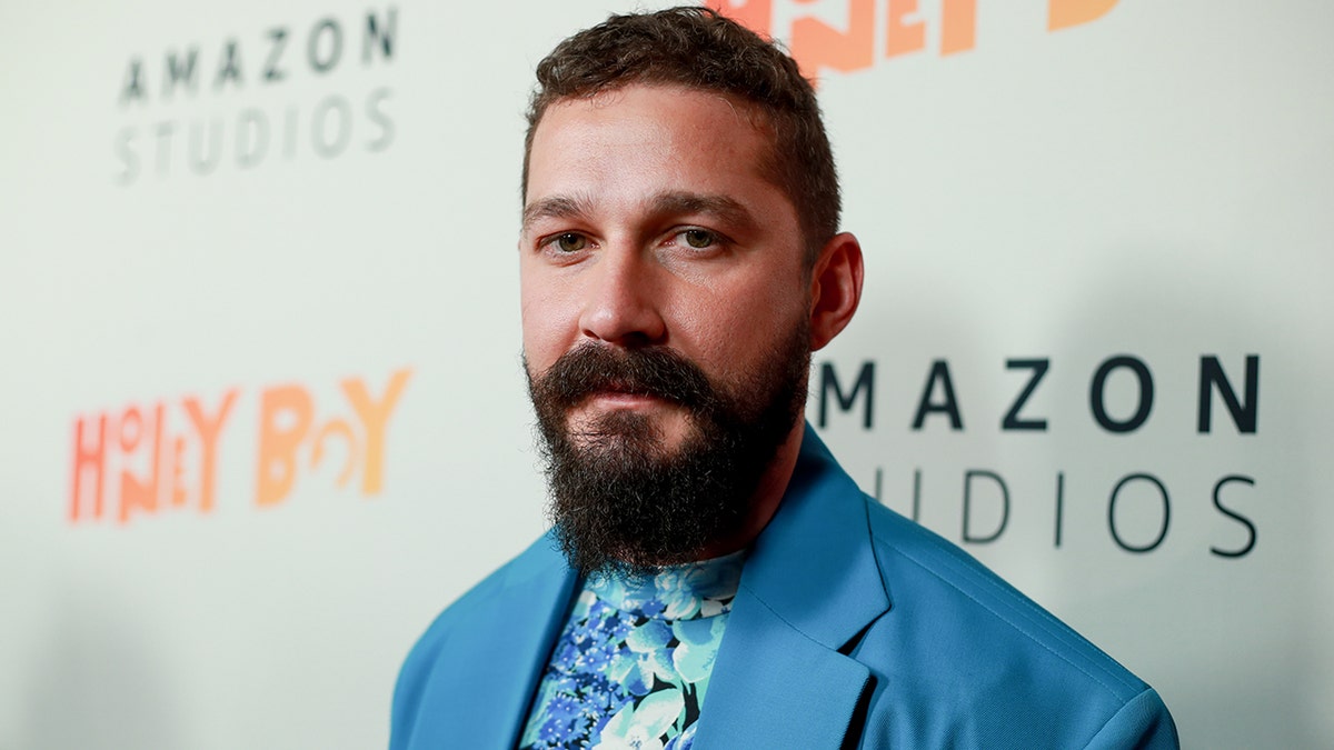 Shia LaBeouf in a blue suit and patterned shirt looks at the camera on the carpet