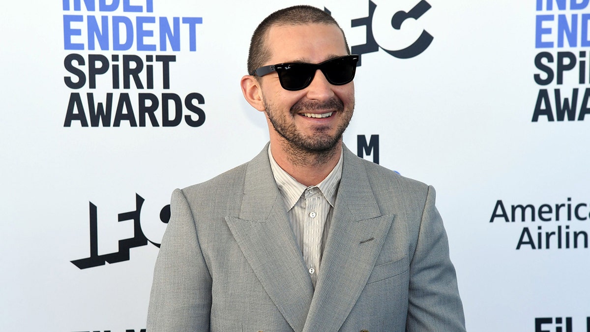 Shia LaBeouf in a grey suit and black sunglasses smiling on the carpet