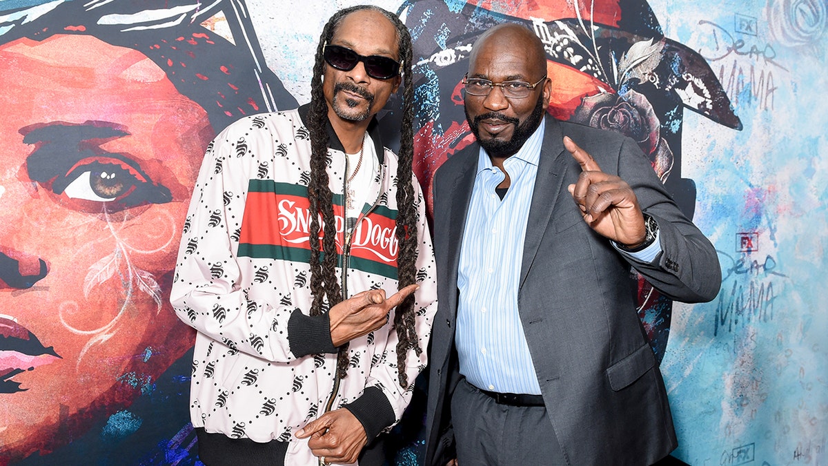 Snoop Dogg com jaqueta branca aponta para Michael "Harry-O" Harris em terno cinza e camisa azul claro