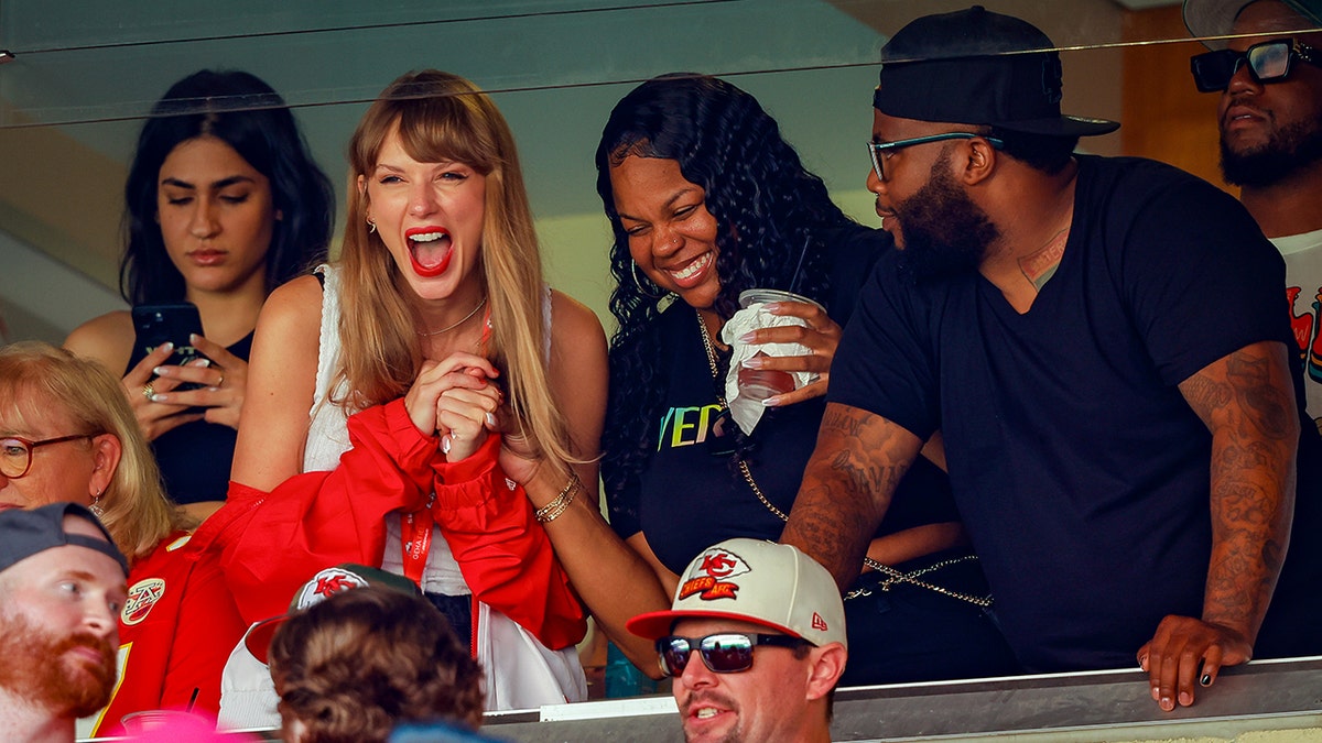 Taylor Swift parece animada em uma blusa branca e jaqueta vermelha enquanto assiste Travis Kelce e os Chiefs jogarem contra os Bears