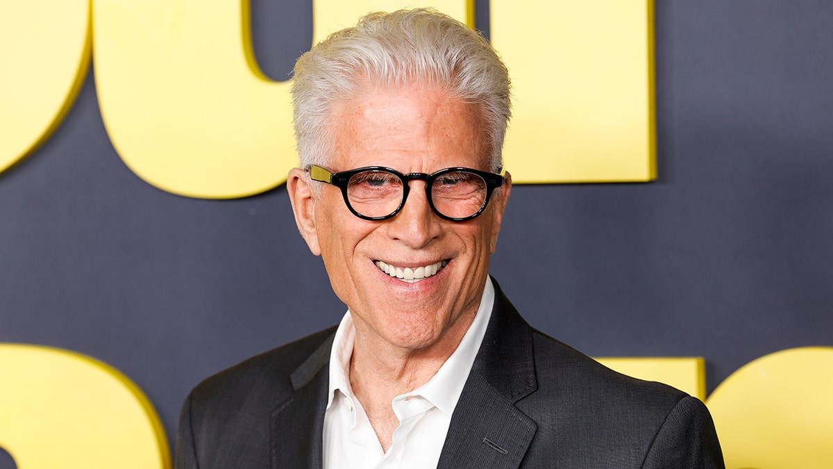 Ted Danson in a white shirt and black blazer with black circular glasses smiles on the carpet