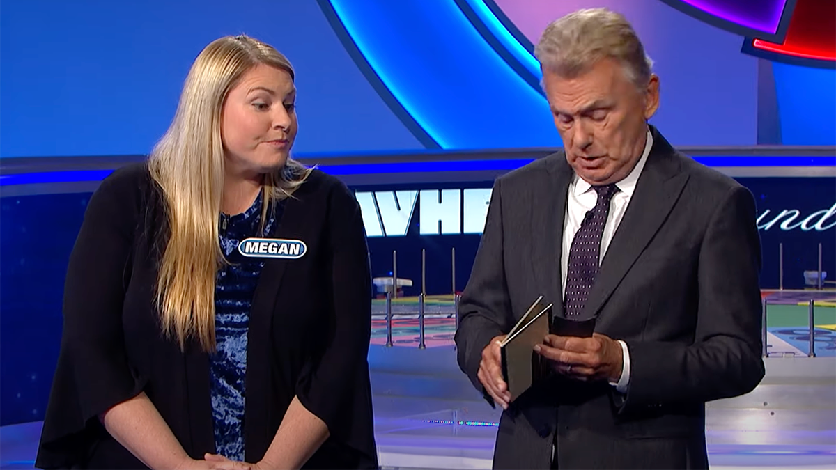 Pat Sajak fumbles with an envelope on the set of "Wheel of Fortune" while contestant Megan looks on