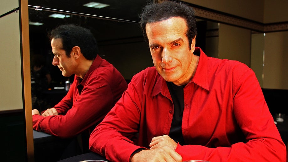 Magician David Copperfield poses in a red shirt for a portrait next to a mirror