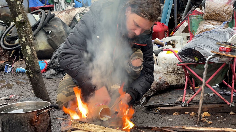 A man tries to light a small campfire in a homeless camp