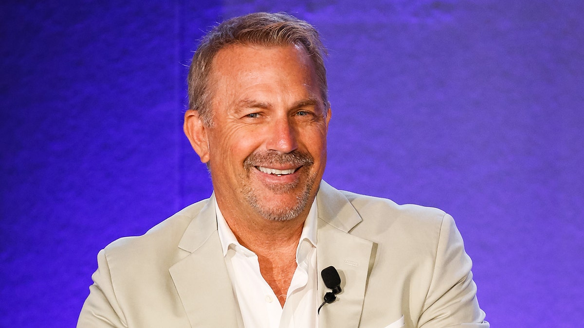 Kevin Costner smiles wearing tan suit and achromatic shirt
