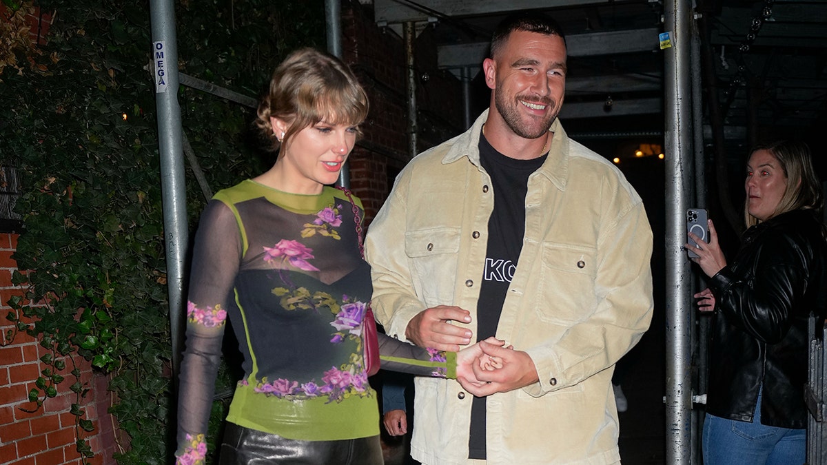 Taylor Swift em um top estampado verde caminha com Travis Kelce em uma jaqueta bege na cidade de Nova York