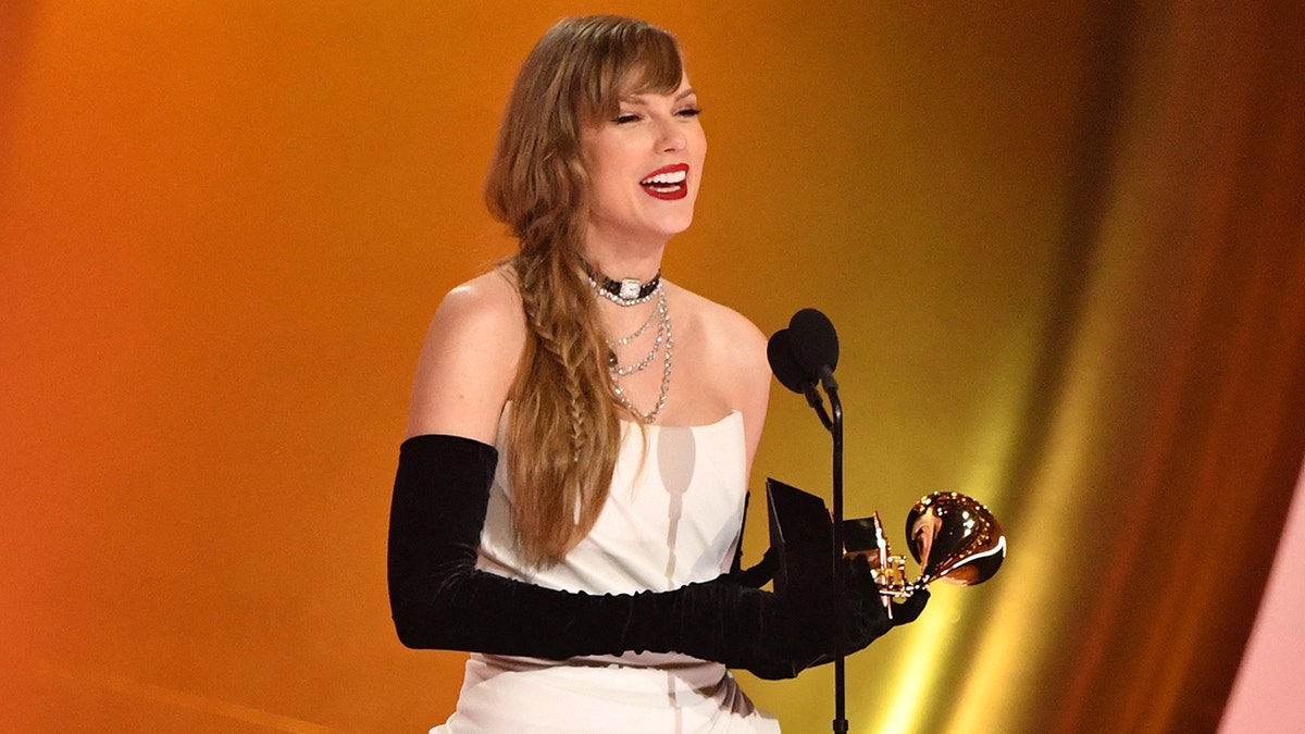 Taylor Swift looks elated on stage at the Grammys holding the award for Best Pop Vocal Album in a white dress with long black gloves