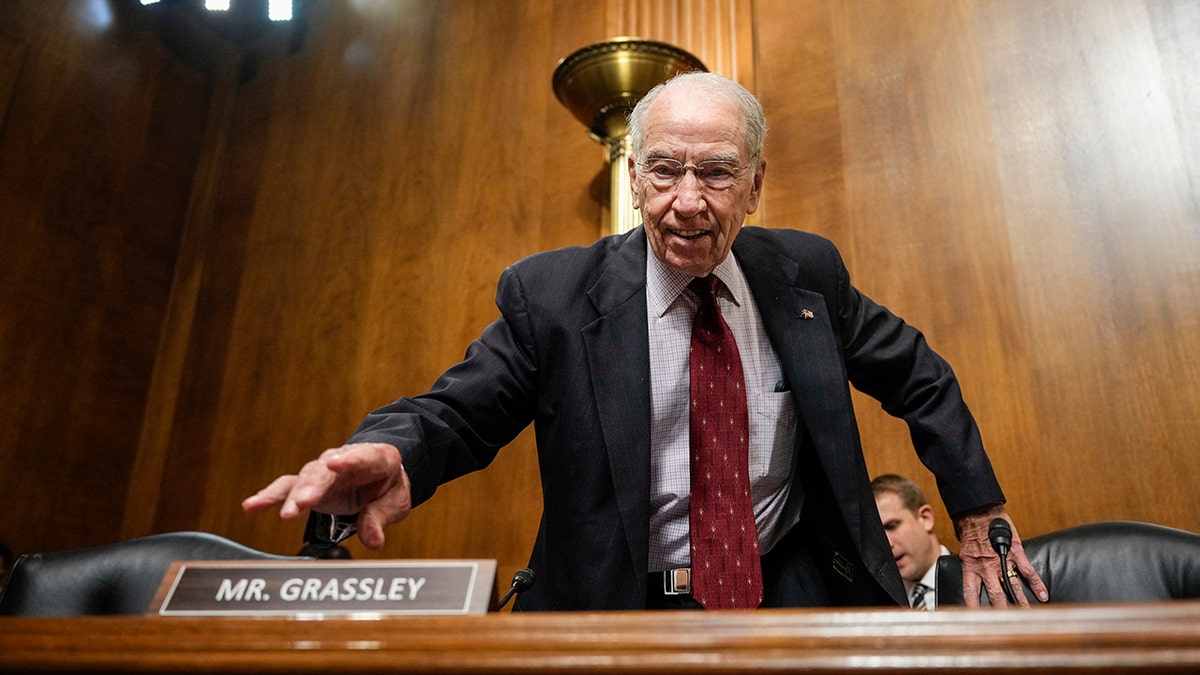 Chuck Grassley successful a acheronian suit, reddish tie, reaching retired correct manus connected table, near manus connected chair