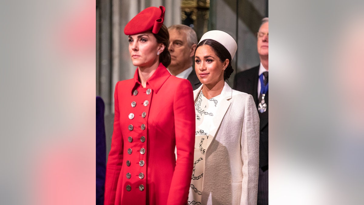 Kate Middleton wearing a red coat dress standing in front of Meghan Markle wearing a white coat drress
