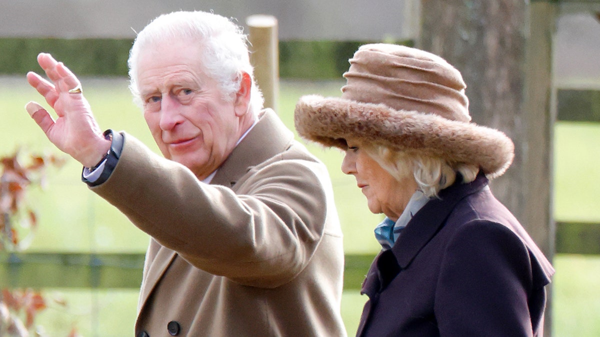 A close-up of King Charles waving outside next to Queen Camilla looking away