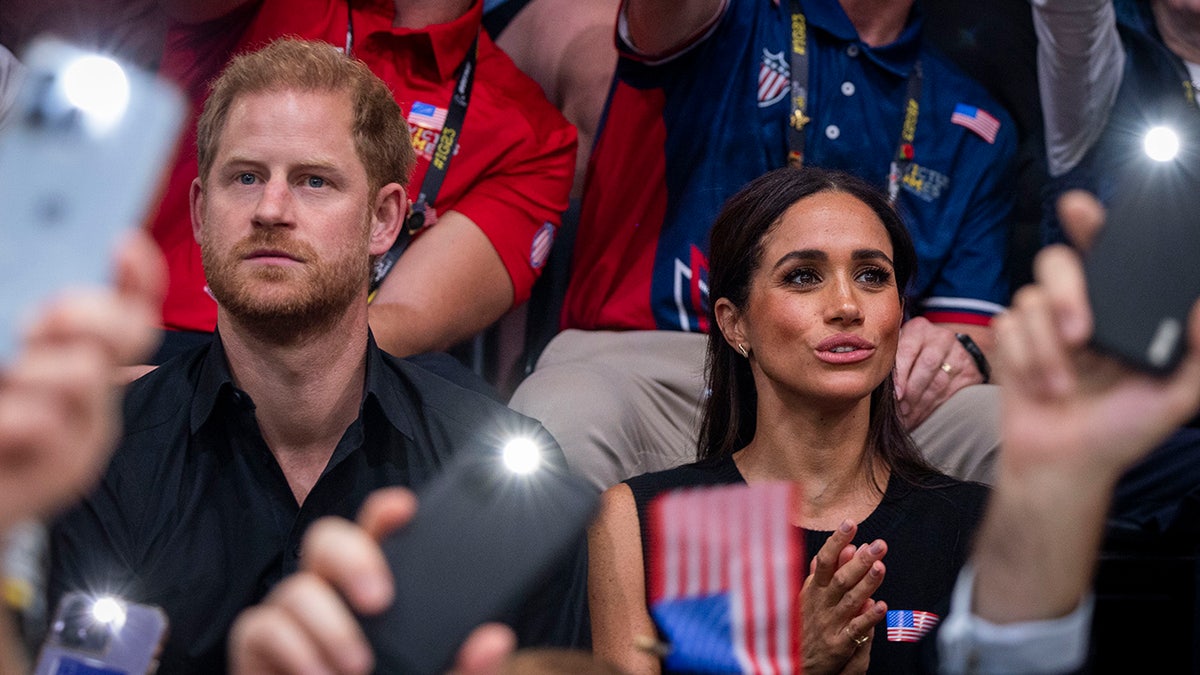 Prince Harry and Meghan Markle sitting in a crowd surrounded by phones
