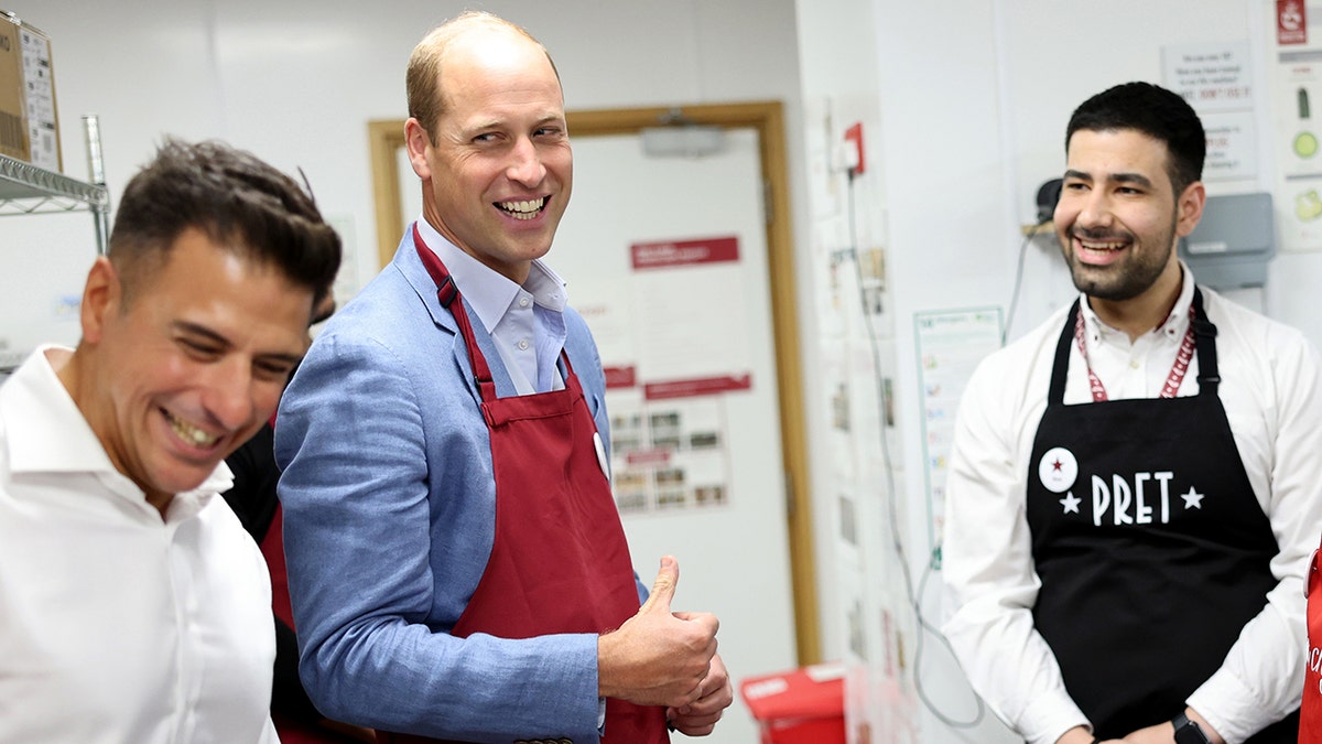 Prince William in a kitchen talking to two men and wearing an apron