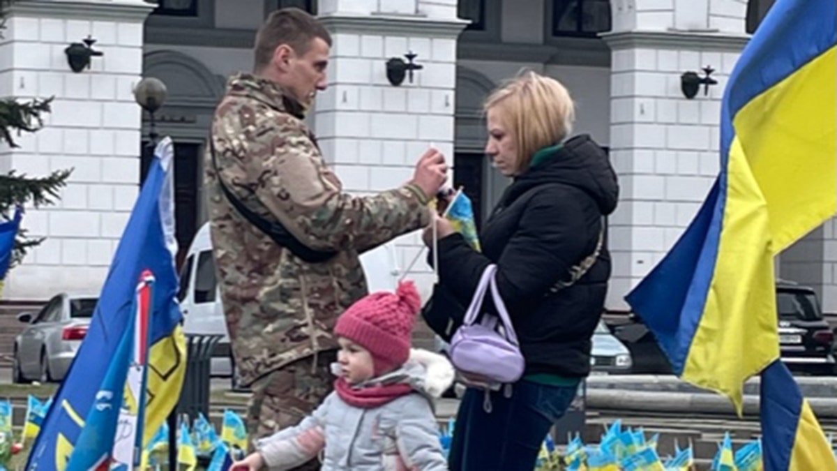 Man in military uniform stands with woman next to a small child surrounded by the flag memorial in Kyiv