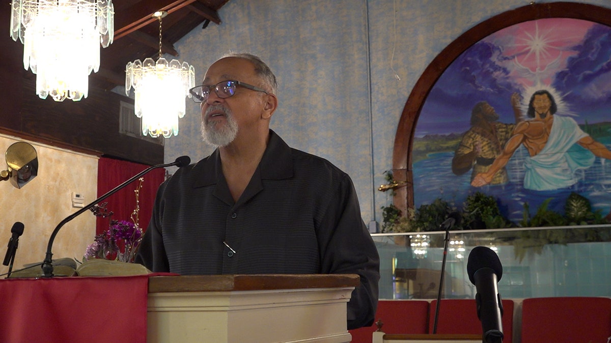 Le pasteur Landry se tient devant l'église en train de lire un passage à sa congrégation