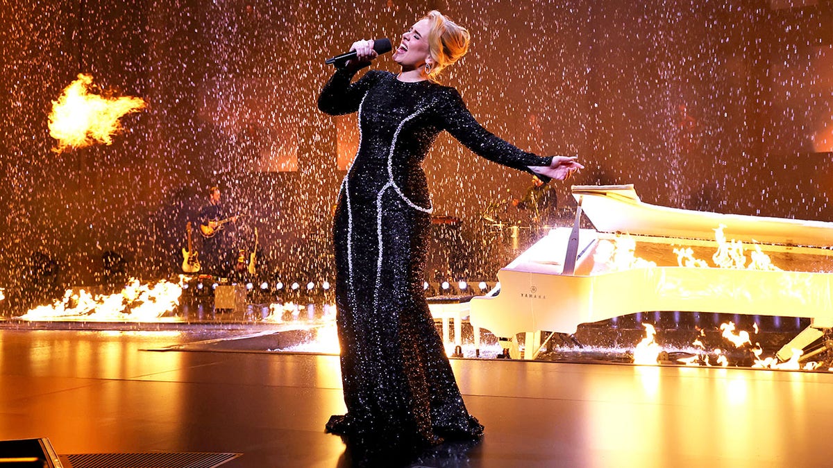 Adele leans back in a black dress singing on stage as fire and rain display in the background