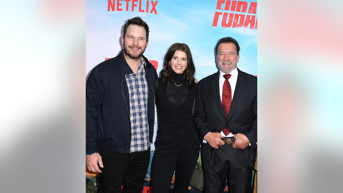 Chris Pratt in a checkered shirt and jacket poses next to wife Katherine in black and her father Arnold in a black suit and red tie
