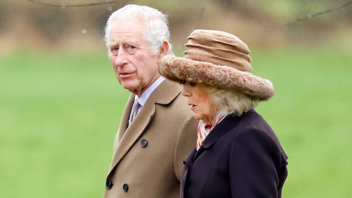 O rei Charles com um casaco bege caminha ao lado da esposa, a rainha Camilla, com um casaco escuro e chapéu bege