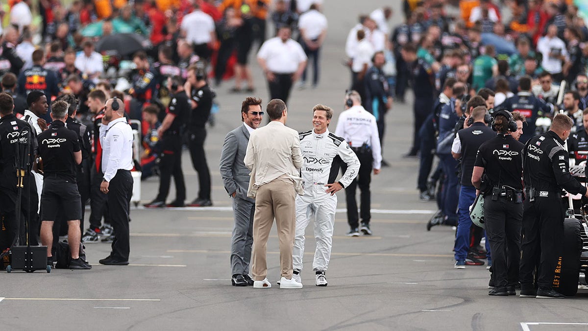 Javier Bardem in a grey suit and Brad Pitt in a white F1 outfit speak with someone on the track with many people on either side of them at the F1 Grand Prix of Great Britain at Silverstone Circuit