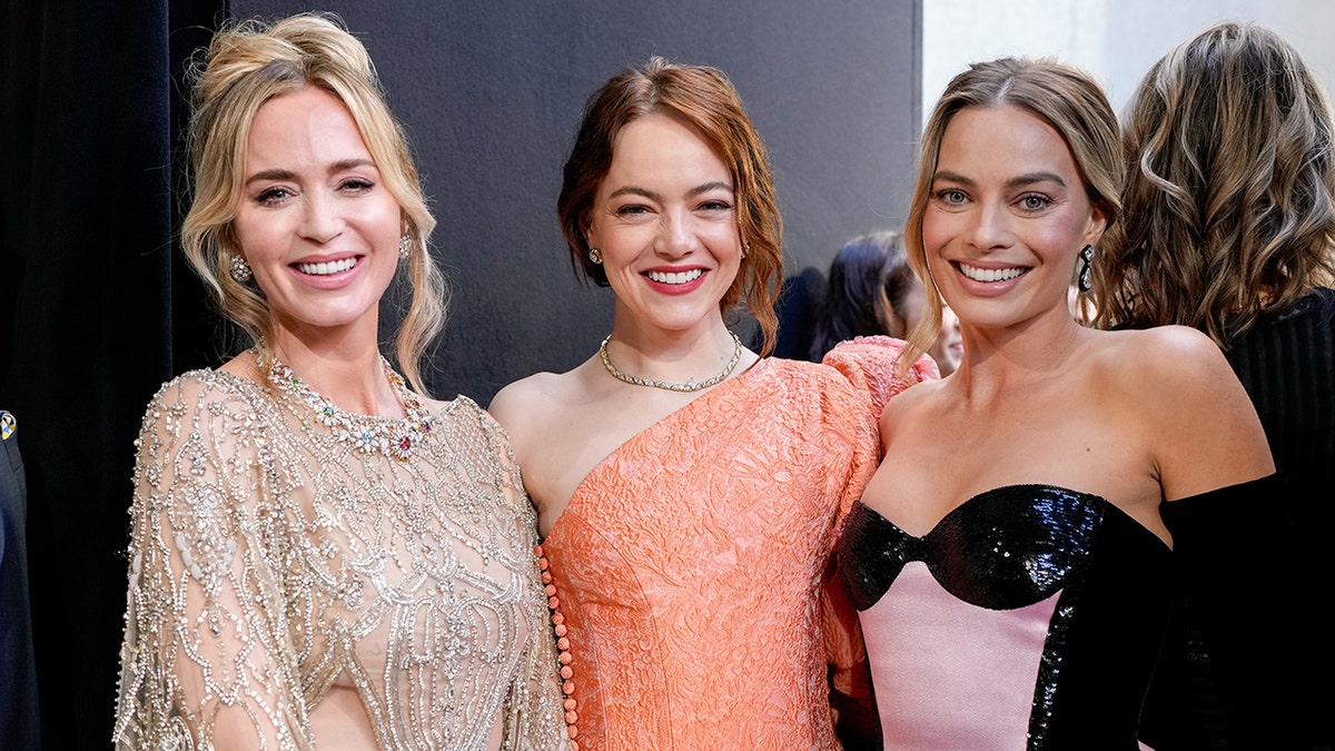 Emily Blunt in a sparkly cut out dress stands next to Emma Stone in an orange sparkly dress who stands next to Margot Robbie in a black and pink dress