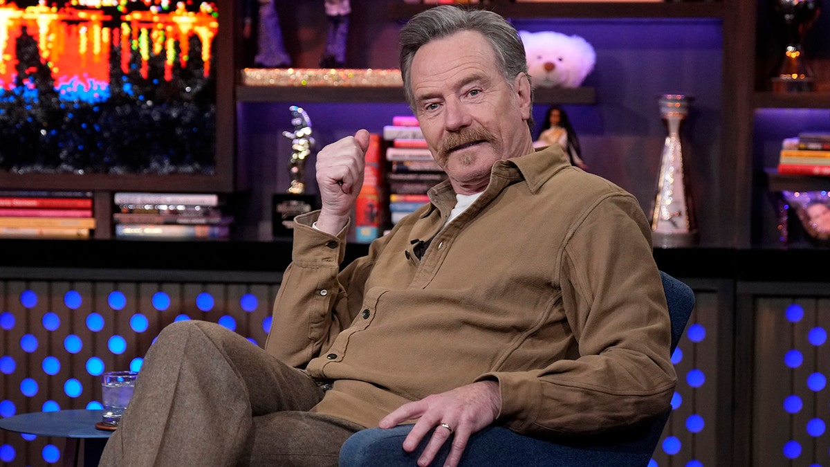 Bryan Cranston in a dark tan sweater sits on a chair at "Watch What Happens Live"
