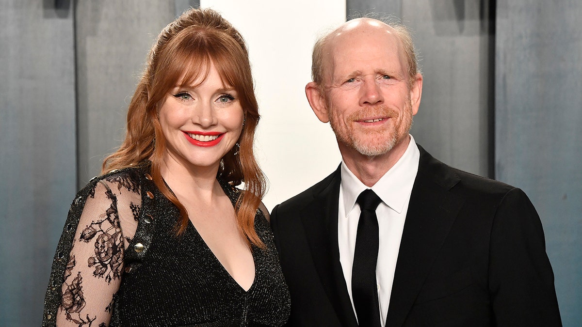Bryce Dallas Howard in a black dress smiles next to her father Ron Howard in a black suit and tie