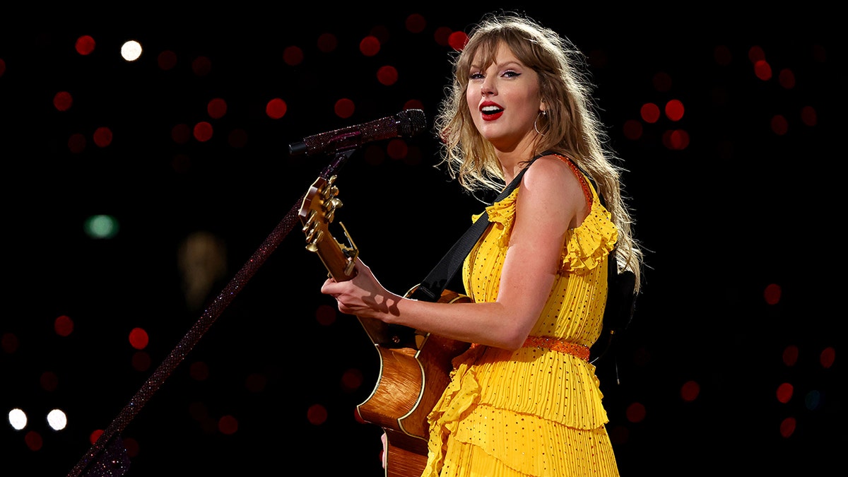 Taylor Swift plays the guitar on stage during her Eras Tour in a bright yellow dress