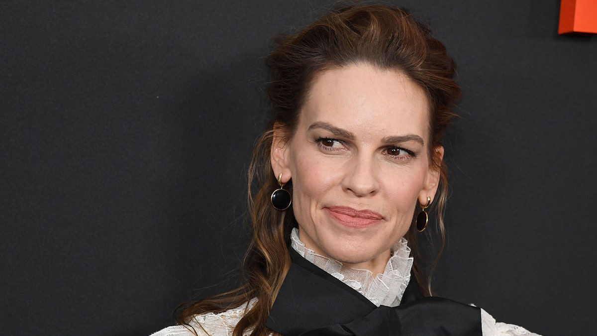 Hilary Swank looks to her right on the carpet in a lace white top with a black bow