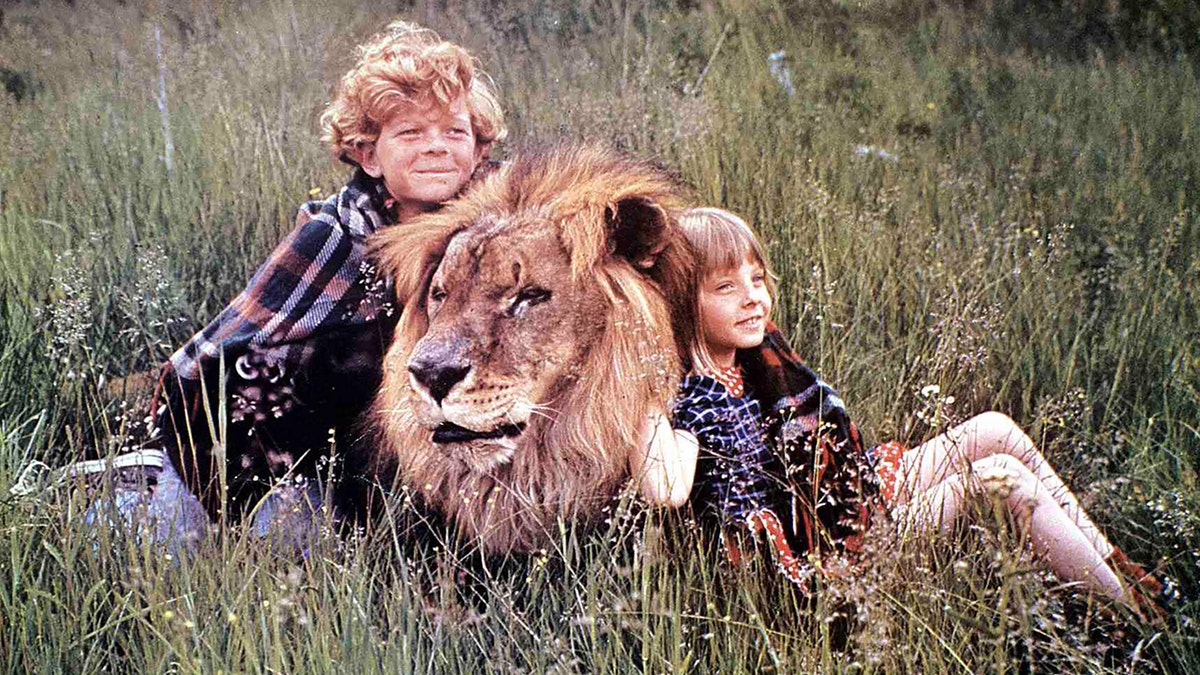 Um menino (Johnny Whitacker) e uma menina (Jodie Foster) sentam-se em um campo com um leão do filme "Napoleão e Samantha"