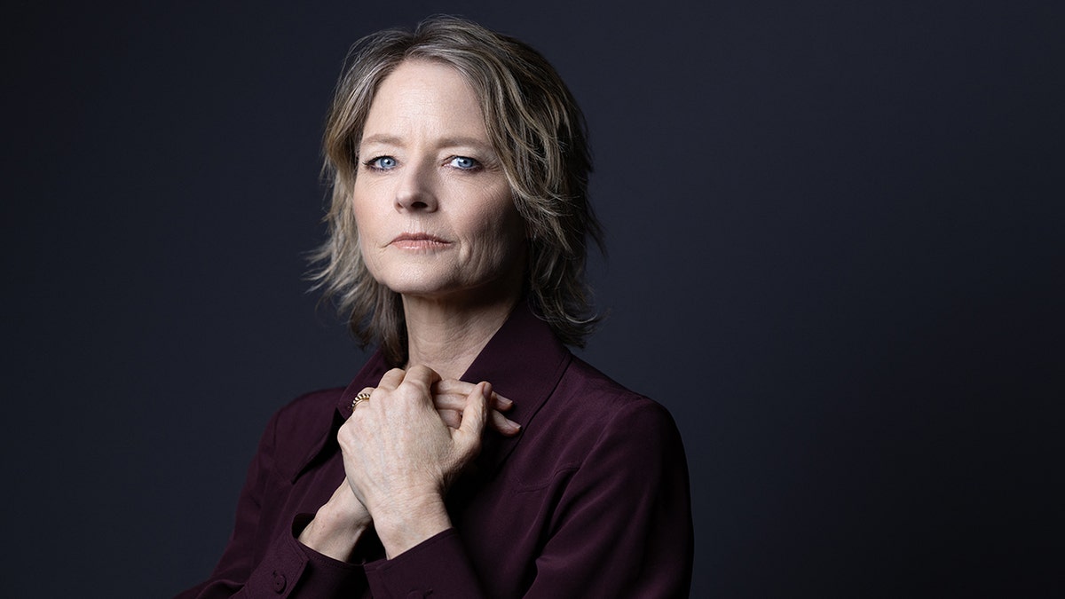 Jodie Foster in a maroon shirt holds her hands to her chest and looks stoic