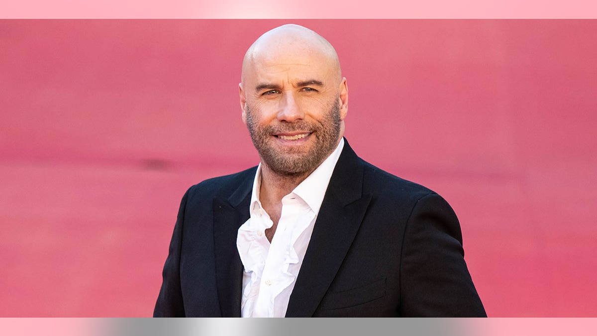 John Travolta in a black suit and white shirt smiles on the carpet