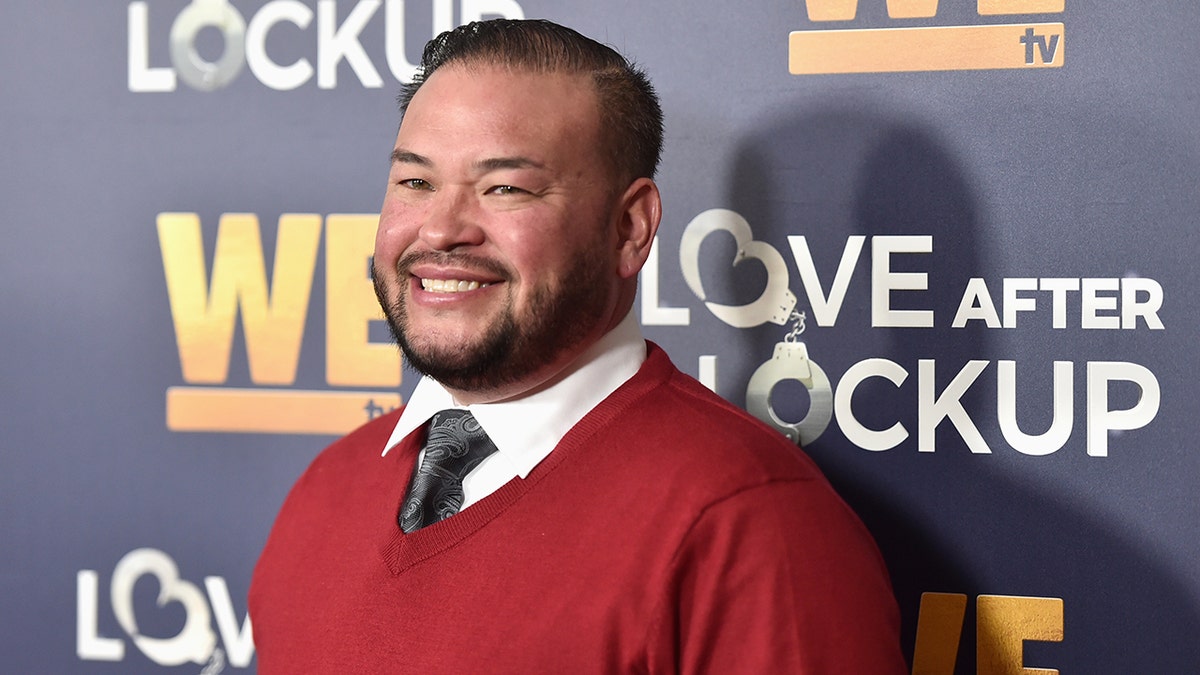 Jon Gosselin in a red sweater and black tie underneath smiles on the carpet