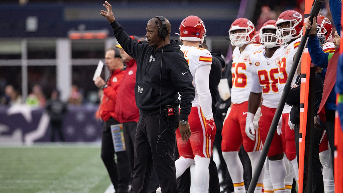 Dave Merritt dressed all in black with headphones and his arm raised while Kansas City Chiefs players stand behind him.