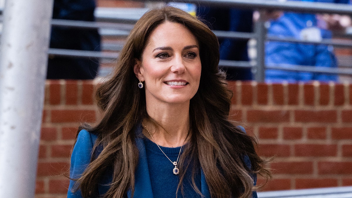 Kate Middleton em uma camisa azul royal e jaqueta combinando caminha e sorri parcialmente