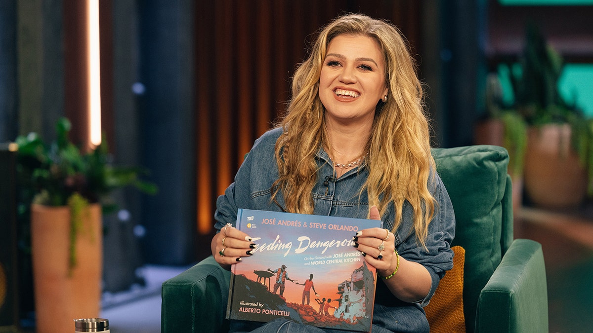 Kelly Clarkson in a jean outfit holds an album and smiles on set of her talk show