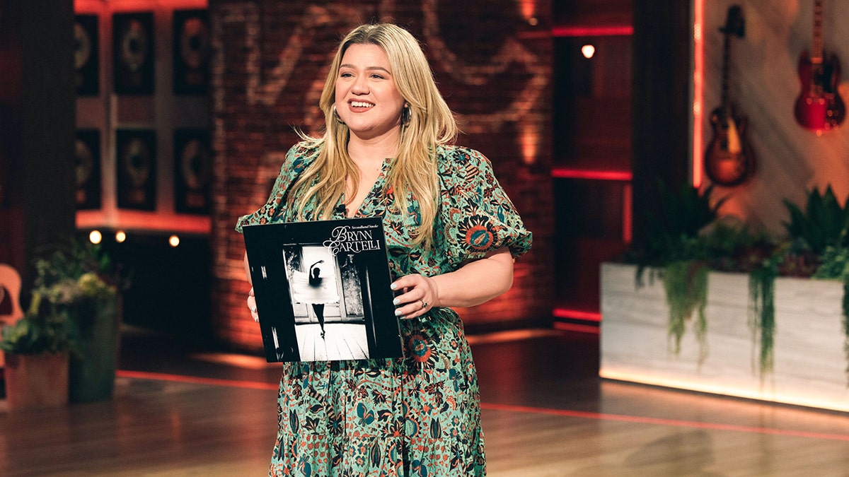 Kelly Clarkson in a green patterned dress holds an album on set of her tv show