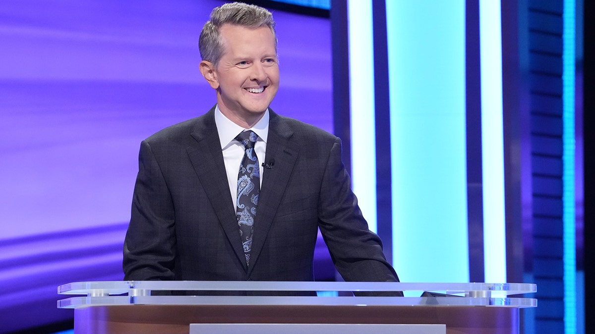 Ken Jennings behind the podium in a black suit and paterned tie for "Jeopardy!"