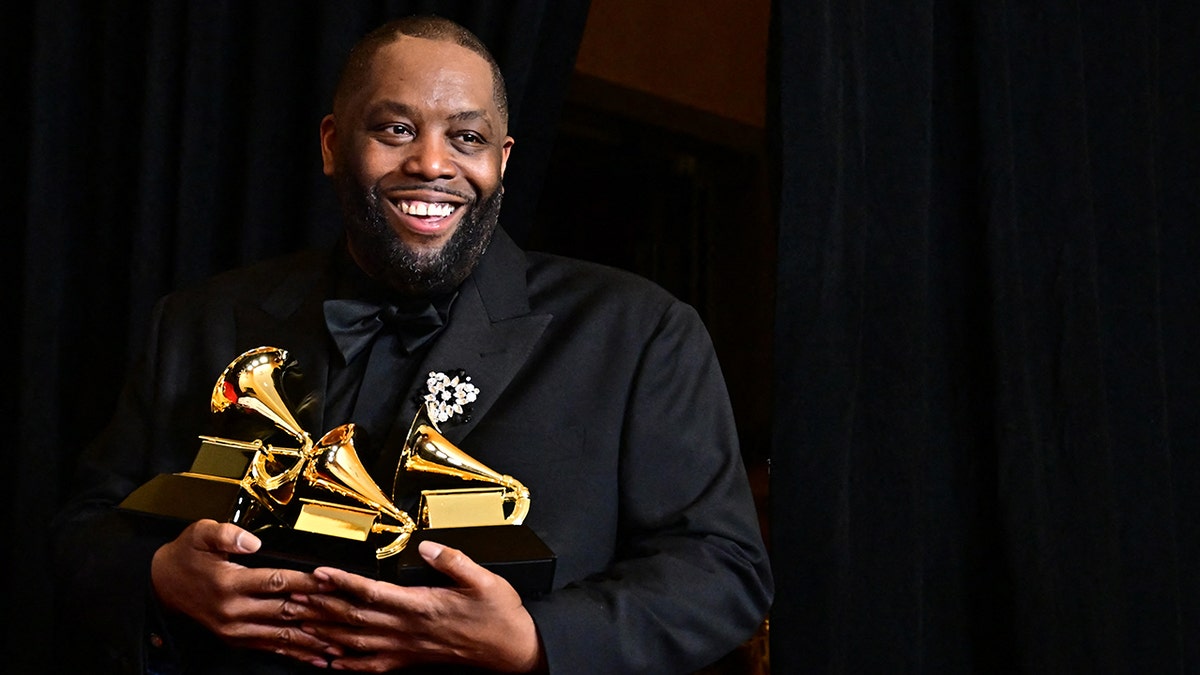 Killer Mike smiles in a black suit holding three Grammy trophies