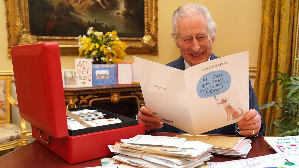 King Charles sits behind his desk and laughs as he reads a card that says "At least you dont have to wear a cone" with a dog in a cone illustrated