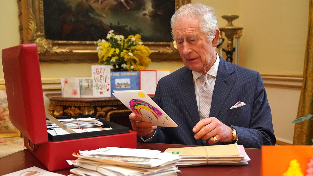 King Charles looks down at a colored card as he sits behind his desk