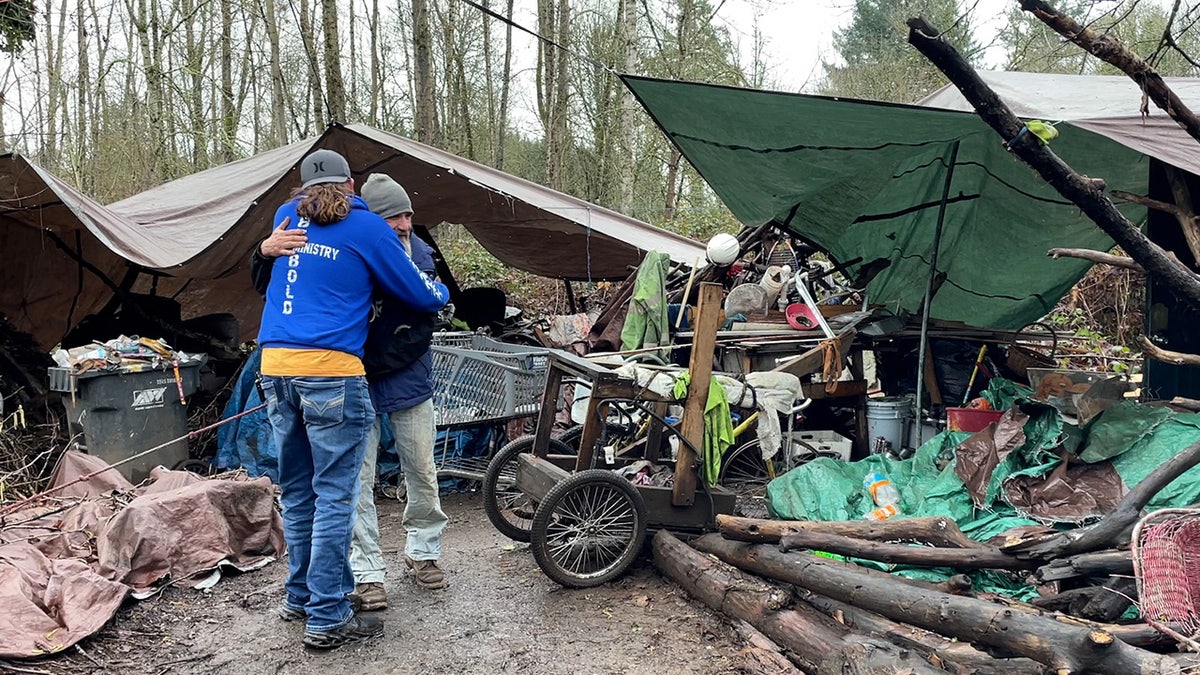 Matt Maceira hugs a friend in front of makeshift shelter in the woods