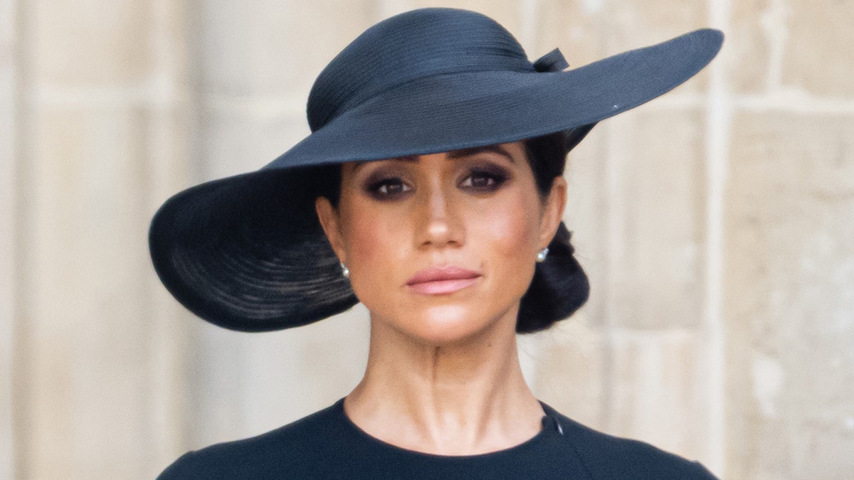 A solemn looking Meghan Markle in black with a tilted black hat