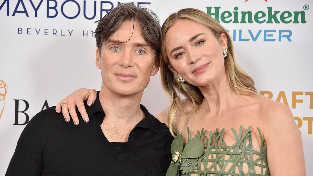 Emily Blunt in a green dress wraps her arm around and leans her head against Cillian Murphy in black on the carpet