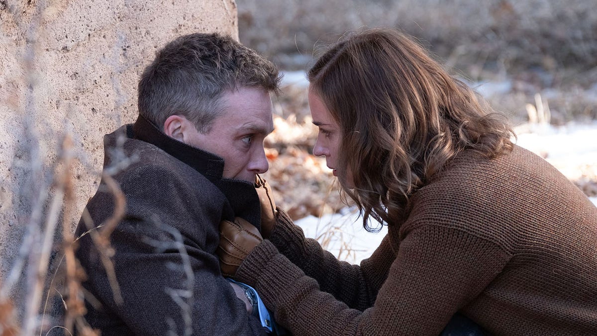 Emily Blunt in a brown jacket grabs onto Cillian Murphy leaning against a tree in a scene from "Oppenheimer"