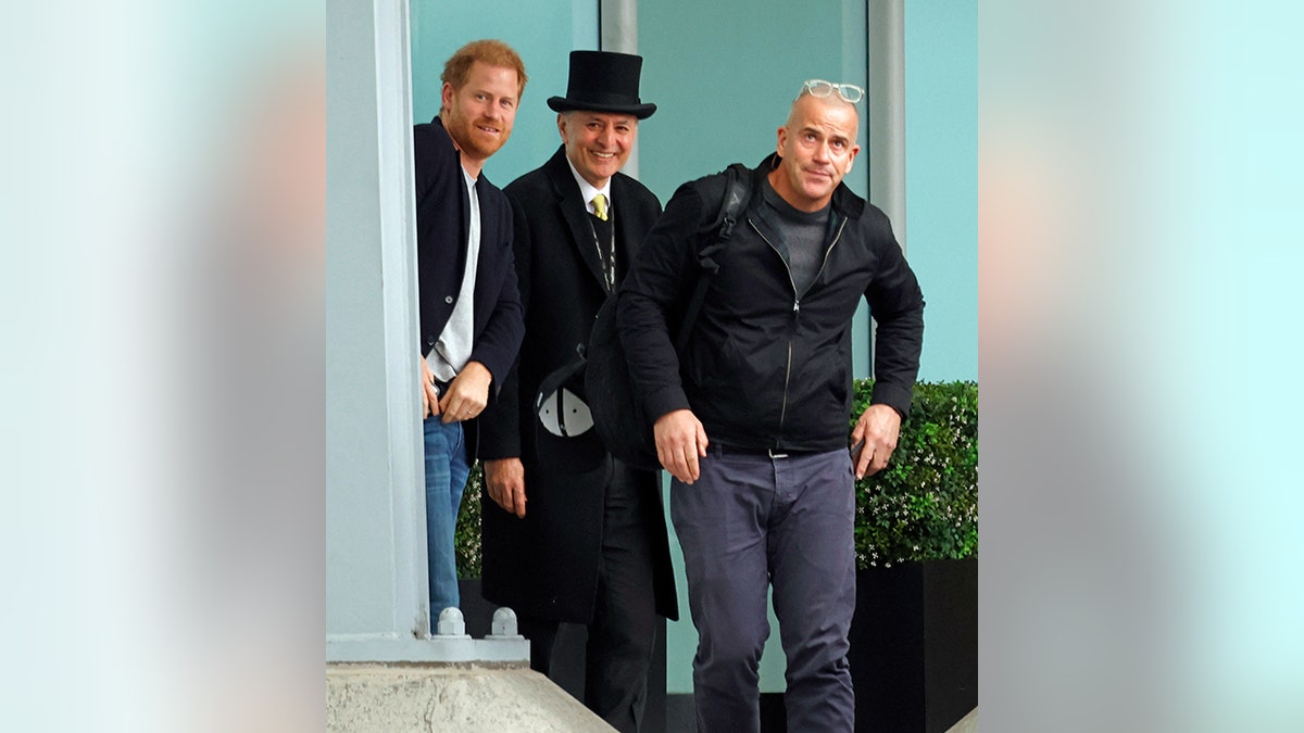 Prince Harry in blue jeans and jacket stands next to a man in a long black hot and top cap and presumably security