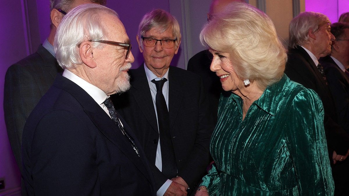 Brian Cox shakes hands with Queen Camilla who is wearing a velvet green dress