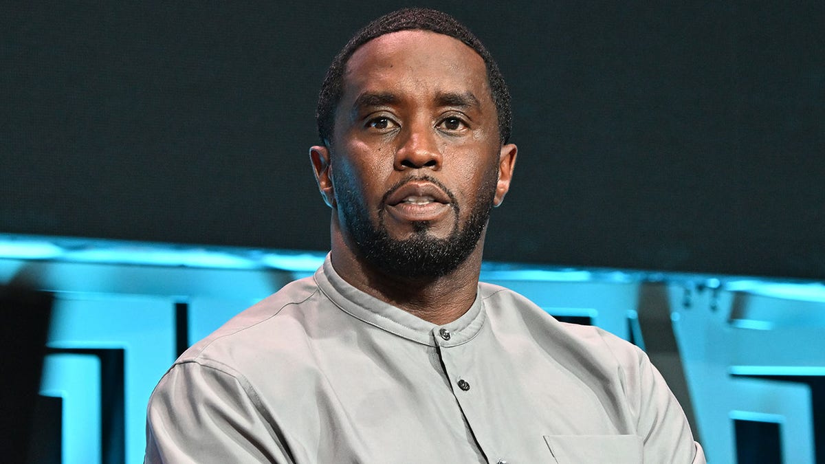 Sean Combs is sitting on stage in a gray shirt and appears immersed in a conversation.