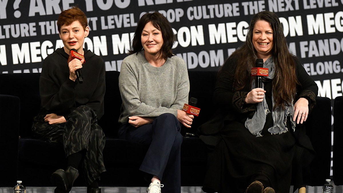 Rose McGowan in black sits next to Shannen Doherty in a grey sweater in the middle, next to Holly Marie Combs in black