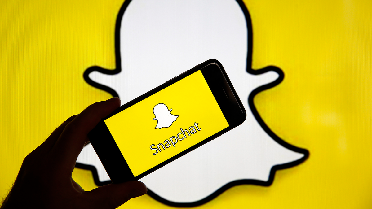 a silhouetted hand holds a phone with the snapchat app loading screen over a snapchat logo