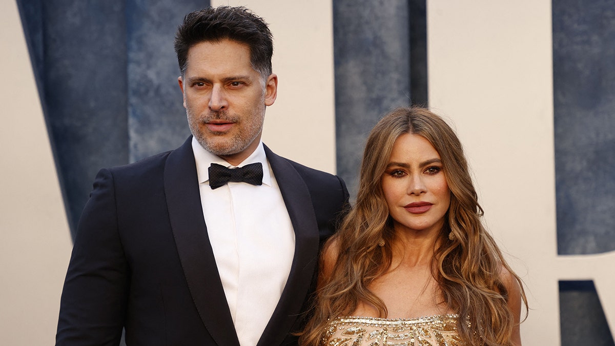 Joe Manganiello in a tuxedo and wife Sofia Vergara in a gold dress with jewels look in opposite directions on the carpet at the Vanity Fair Oscar Party