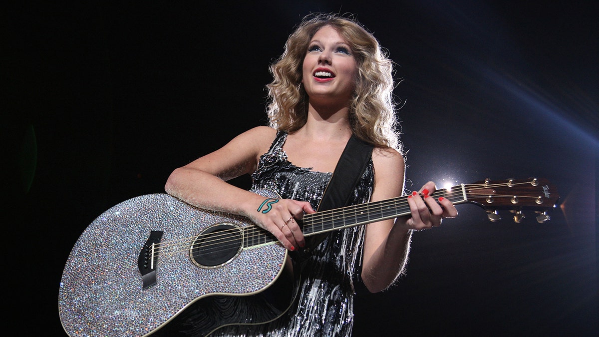 Taylor Swift in a sparkly dress plays on a bejeweled guitar with the number 13 written on her hand
